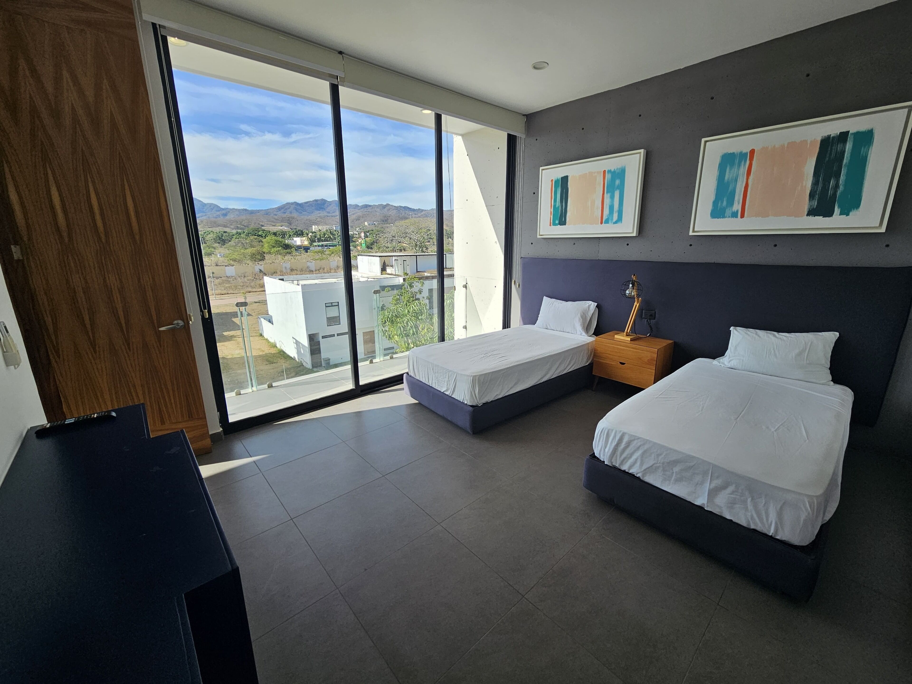 A modern hotel room with two beds, a desk, and a large window offering a view of the mountains.
