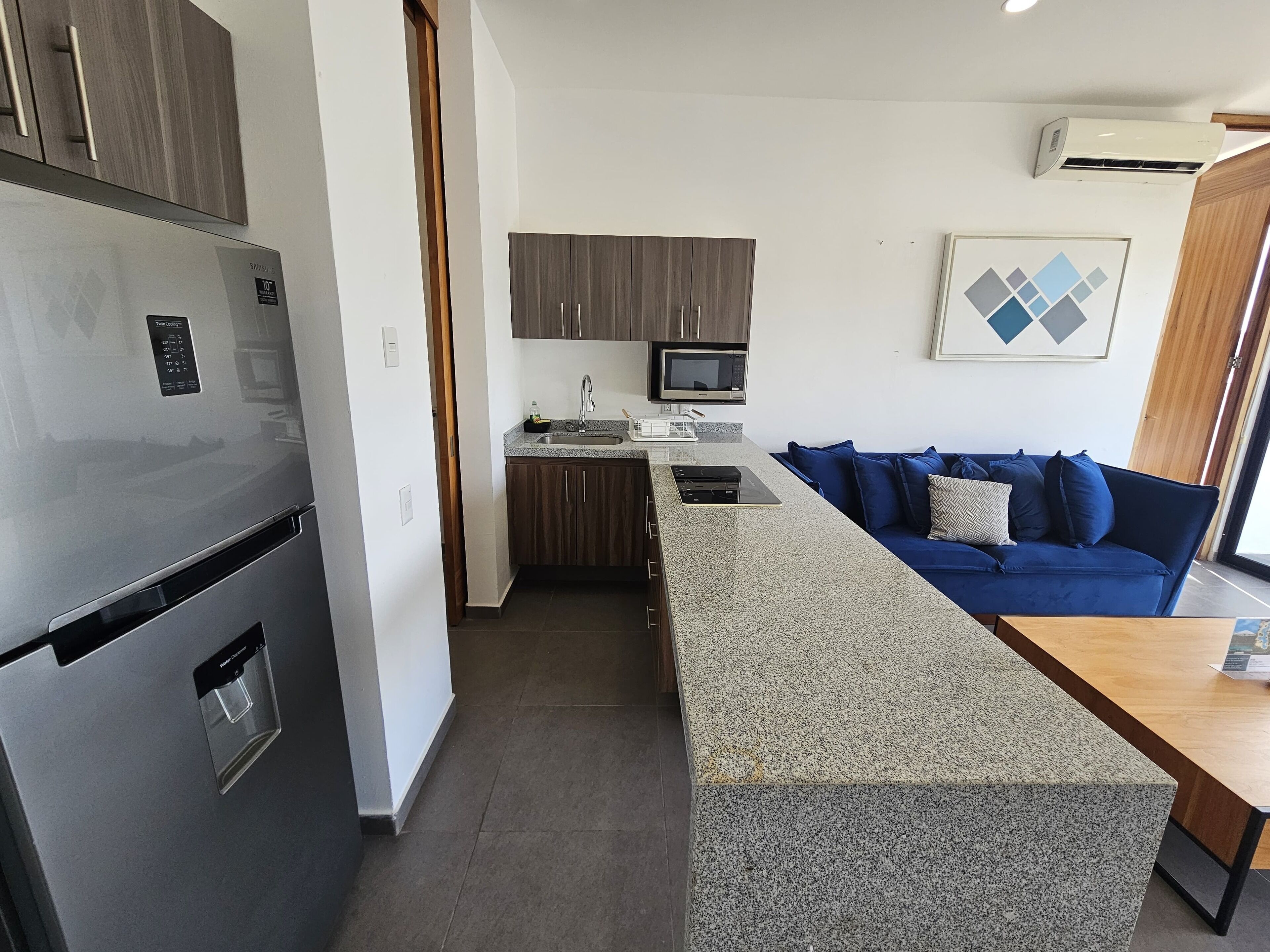 A compact kitchen with a built-in microwave, a refrigerator, and a sink. There is a blue sofa and a wooden table in the living area.