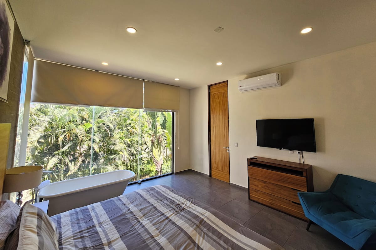 Une chambre moderne avec une grande fenêtre, un lit, un banc, une télévision et une commode.
