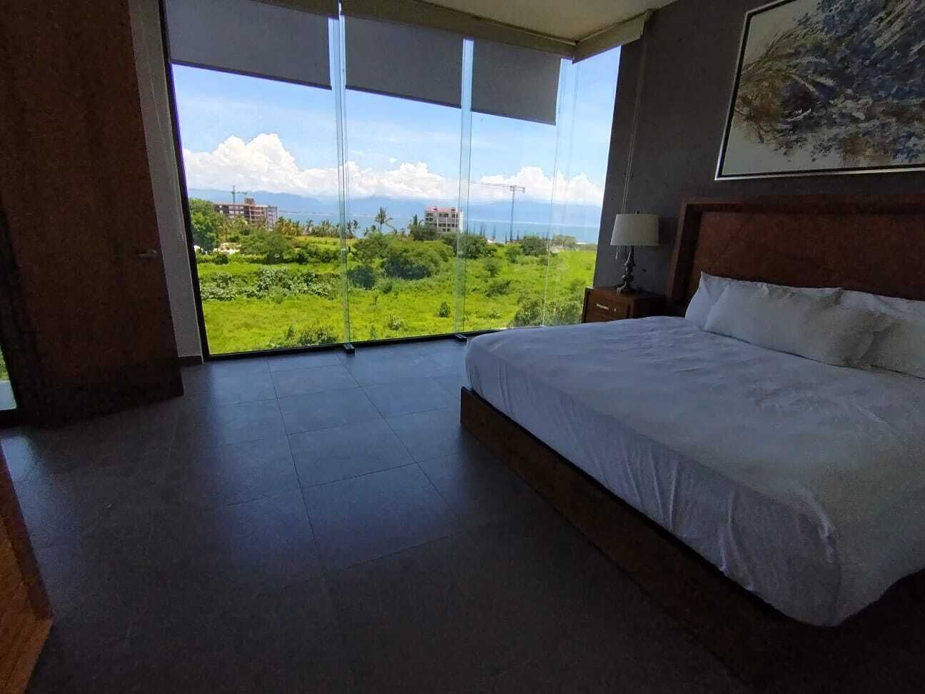 A bedroom with a large bed, a nightstand, and a view of a green landscape through a glass door.