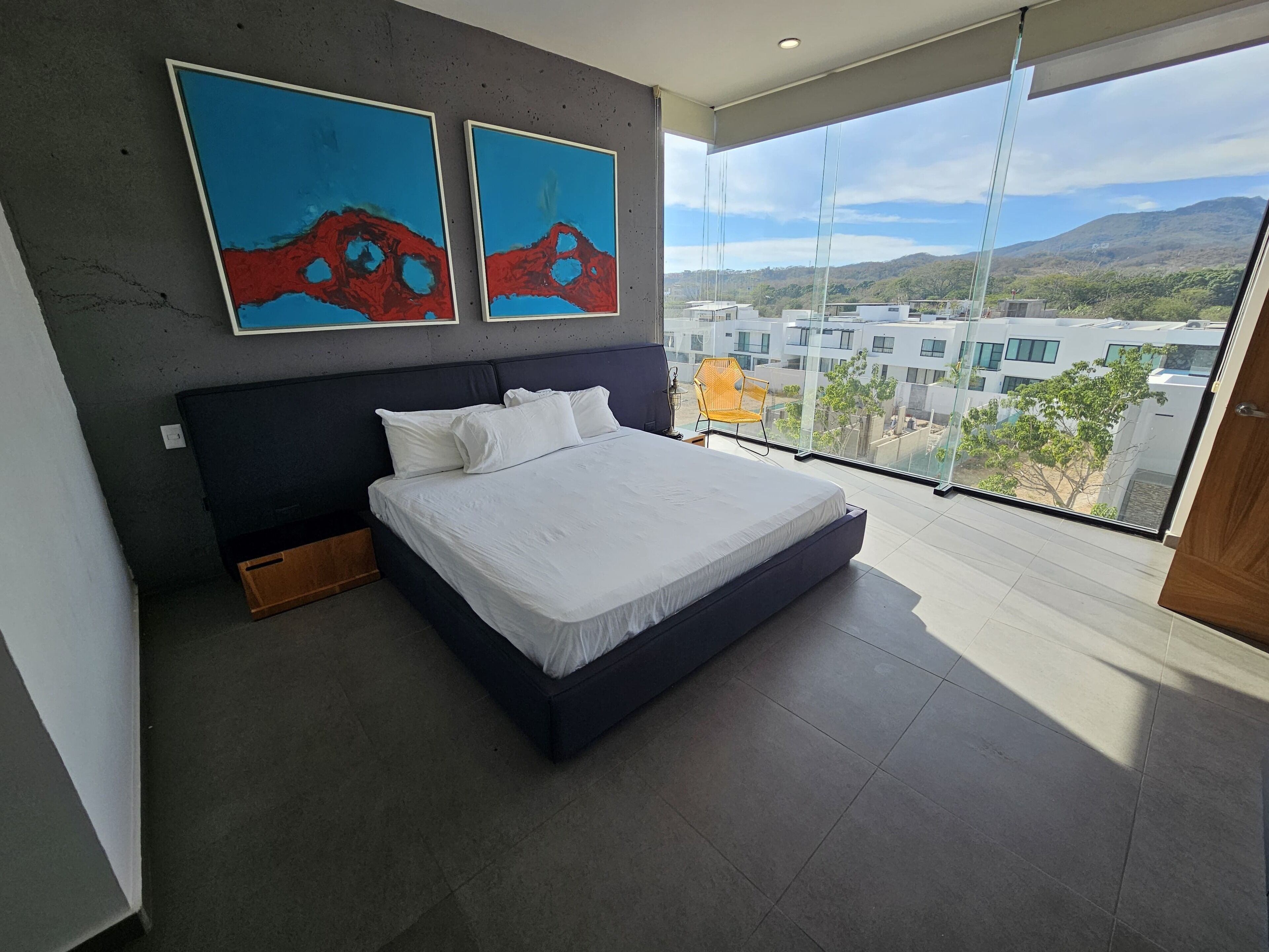 Un dormitorio moderno con una cama grande, dos cuadros abstractos y vistas a edificios y montañas a través de amplios ventanales.