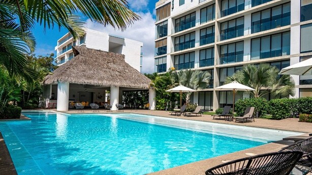A swimming pool with lounge chairs and umbrellas, a thatched-roof structure, and a modern building with balconies. - Torres Mittah (La Cruz de Huanacaxtle)