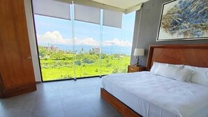 A bedroom with a large bed, a wooden headboard, a nightstand, and a view of a green landscape through floor-to-ceiling windows. - Torres Mittah (La Cruz de Huanacaxtle)
