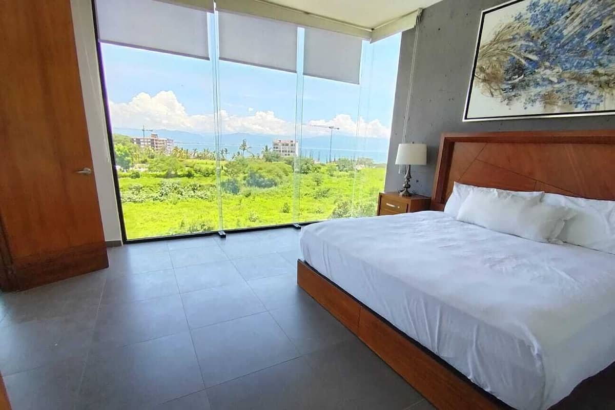 Une chambre dotée d’un grand lit, d’une tête de lit en bois, d’une table de chevet et offrant une vue sur un paysage verdoyant grâce à de grandes fenêtres du sol au plafond.