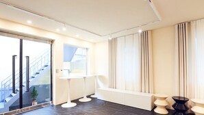 Un lobby moderno de hotel con entrada por puerta de vidrio, una escalera, un área de descanso con mesas redondas y un amplio ventanal con cortinas.