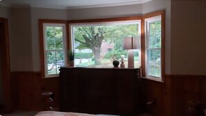 Una habitación con un ventanal grande, un cabecero de madera y una mesita de noche con una lámpara.