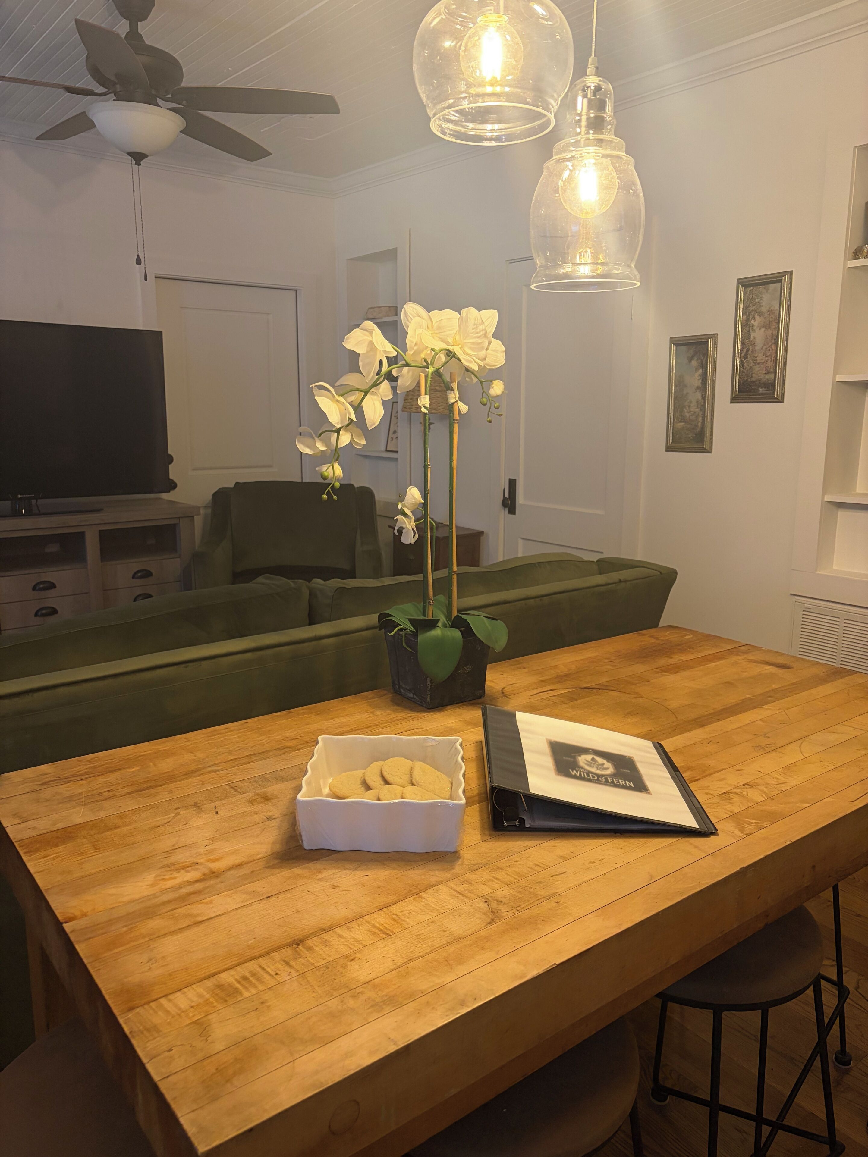 Une table de salle à manger en bois, sur laquelle se trouvent un bol blanc rempli de biscuits, un classeur noir et une orchidée en pot.