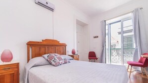 Et soveværelse med træsenggavl, seng med mønstret sengetøj, et natbord med lampe, en stol og en balkon med udsigt.