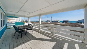 Un espacio al aire libre techado con una mesa, sillas, barandal blanco y vista a la playa y al océano.