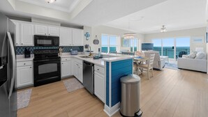 Een moderne keuken met witte kasten, een zwarte kookplaat en een vaatwasser. Er is een eethoek met een tafel en stoelen, en uitzicht op de oceaan door grote ramen.