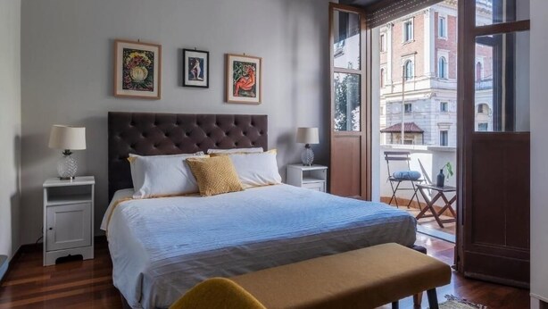 A hotel room with a large bed, two bedside tables, a window with a view of buildings, and three framed artworks on the wall. - HospitalityRome Artists House (Roma)