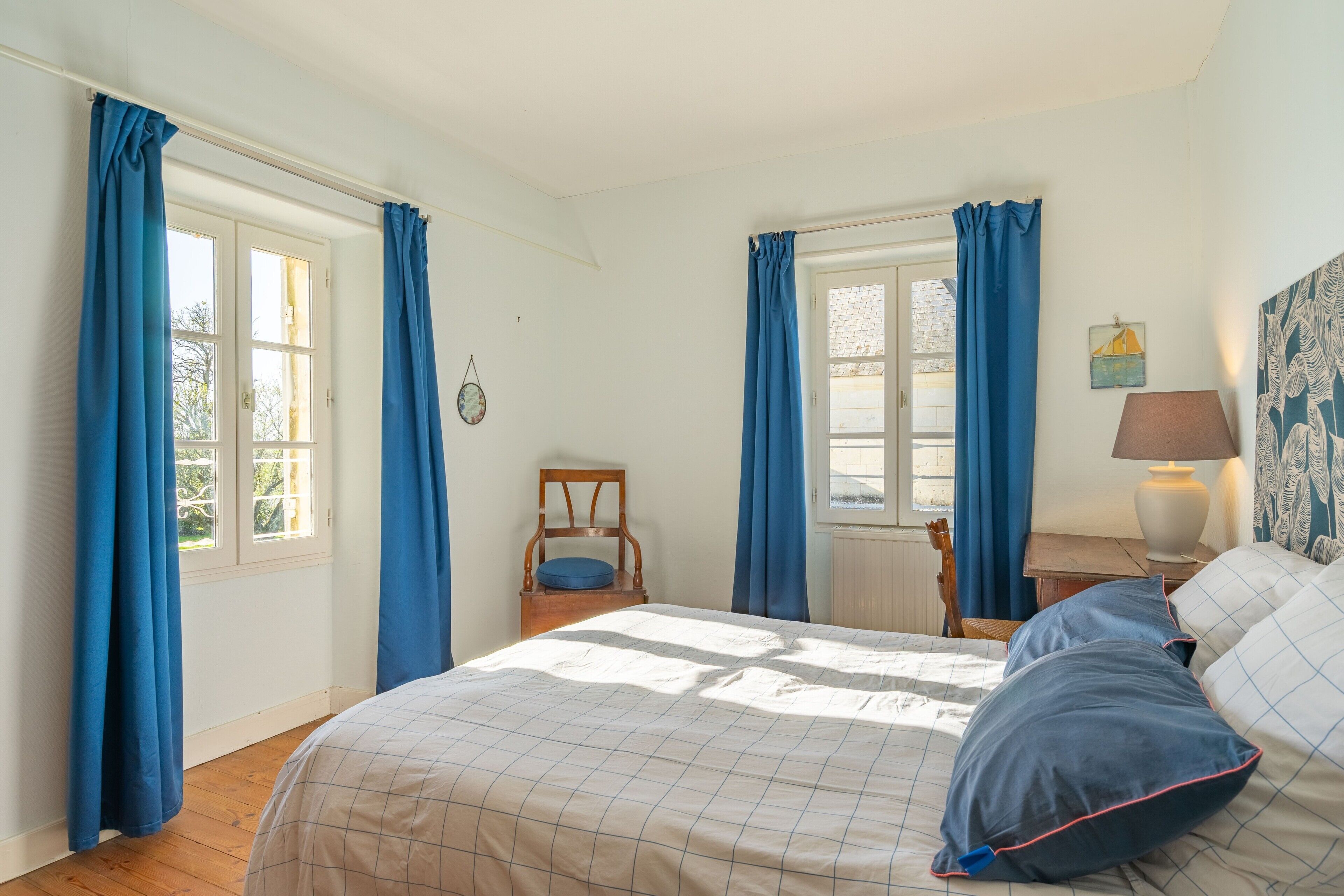A bedroom with a bed, two windows with curtains, a chair, a nightstand with a lamp, and a wall painting.