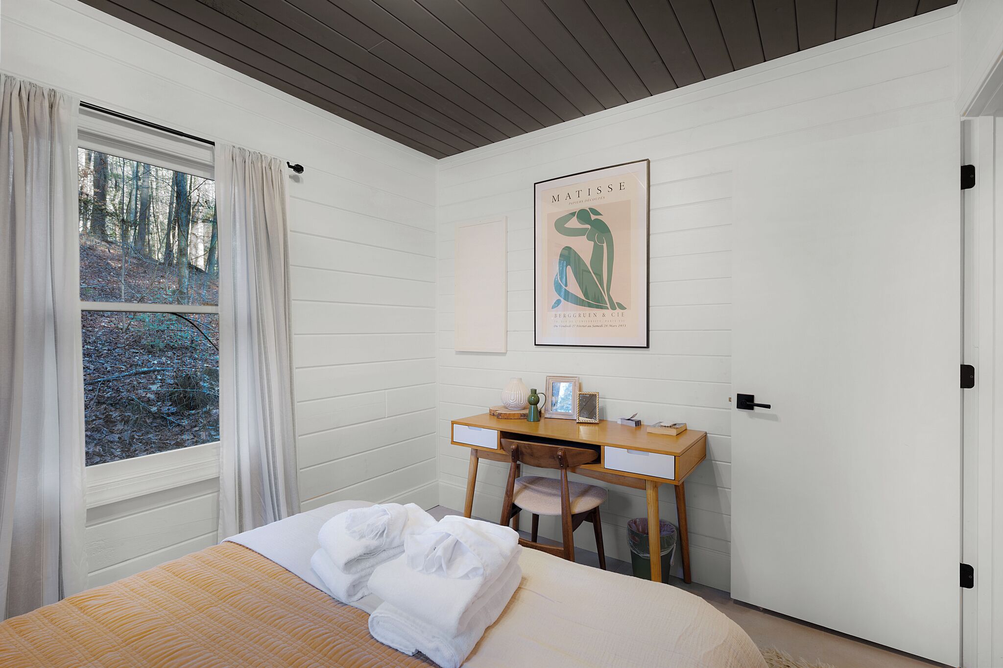 A bedroom with a bed, a desk, and a chair. There is a window with curtains, a framed artwork on the wall, and a door.