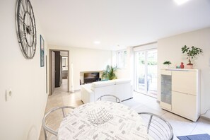 A modern living room with a dining table, a large clock, a TV, and a cabinet with plants. - Le Victor Apartment – Comfortable Stay in Aix-les-Bains (Aix-les-Bains)