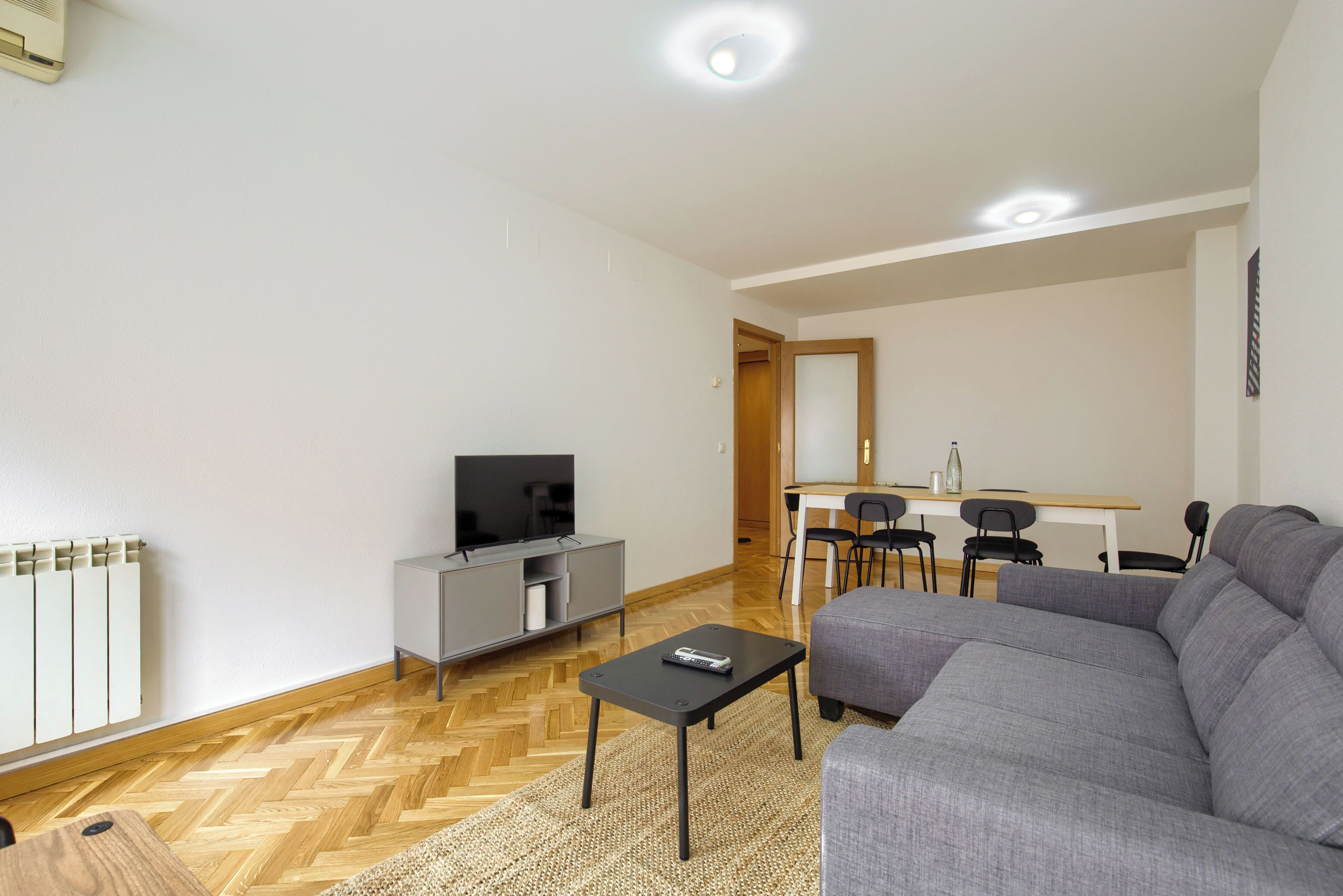 A modern living room with a grey sofa, a small black table, a TV on a grey cabinet, and a dining area with a wooden table and chairs.