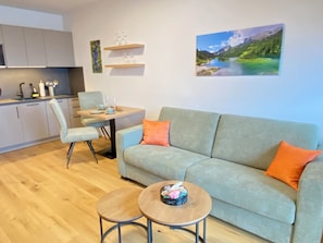 A modern living room with a sofa, coffee table, and dining table. There is a kitchen area with cabinets and a sink. A painting of a landscape is on the wall. - Grimming View I  (Tauplitz)
