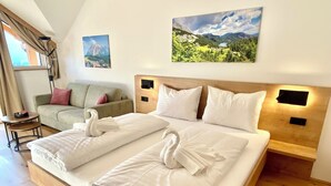 Ein Doppelbett mit weißen Bettwäsche und zwei Handtüchern, ein hölzernes Kopfteil, ein kleiner runder Tisch mit Stuhl, ein Sofa mit Kissen und zwei gerahmte Bilder an der Wand.