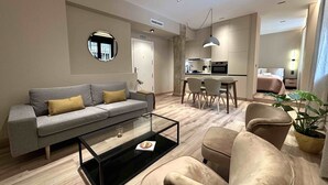 Ein modernes Wohnzimmer mit Sofa, Couchtisch und Sesseln, ein Küchenbereich mit Esstisch und ein Bett im Hintergrund.