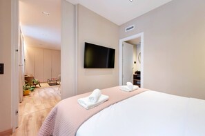 A hotel room with a bed, a flat-screen TV mounted on the wall, a door leading to a bathroom, and a small seating area with a chair and a table. - STAYMADRID - Chic Apartment (MADRID)