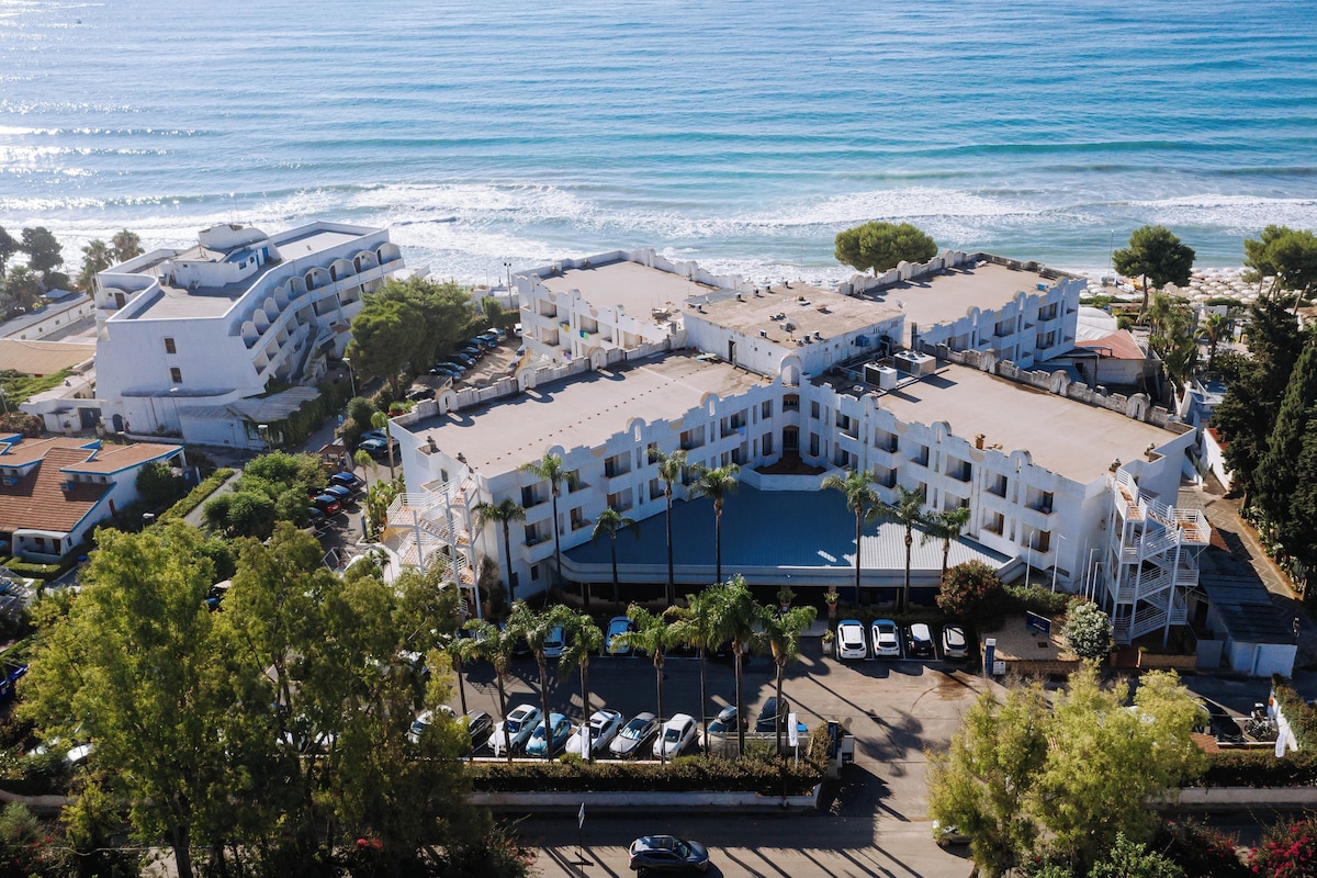 Vista aérea de un hotel costero con varias edificaciones, un estacionamiento y vistas al mar.