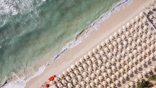 Vista aérea de una playa con hileras de sombrillas y sillas de playa blancas, una orilla arenosa y olas suaves.