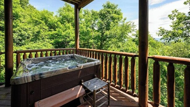 A hot tub on a wooden deck with a wooden railing, surrounded by lush greenery. - Smoky Mountain Bliss by Beyond Expectations (Sevierville)