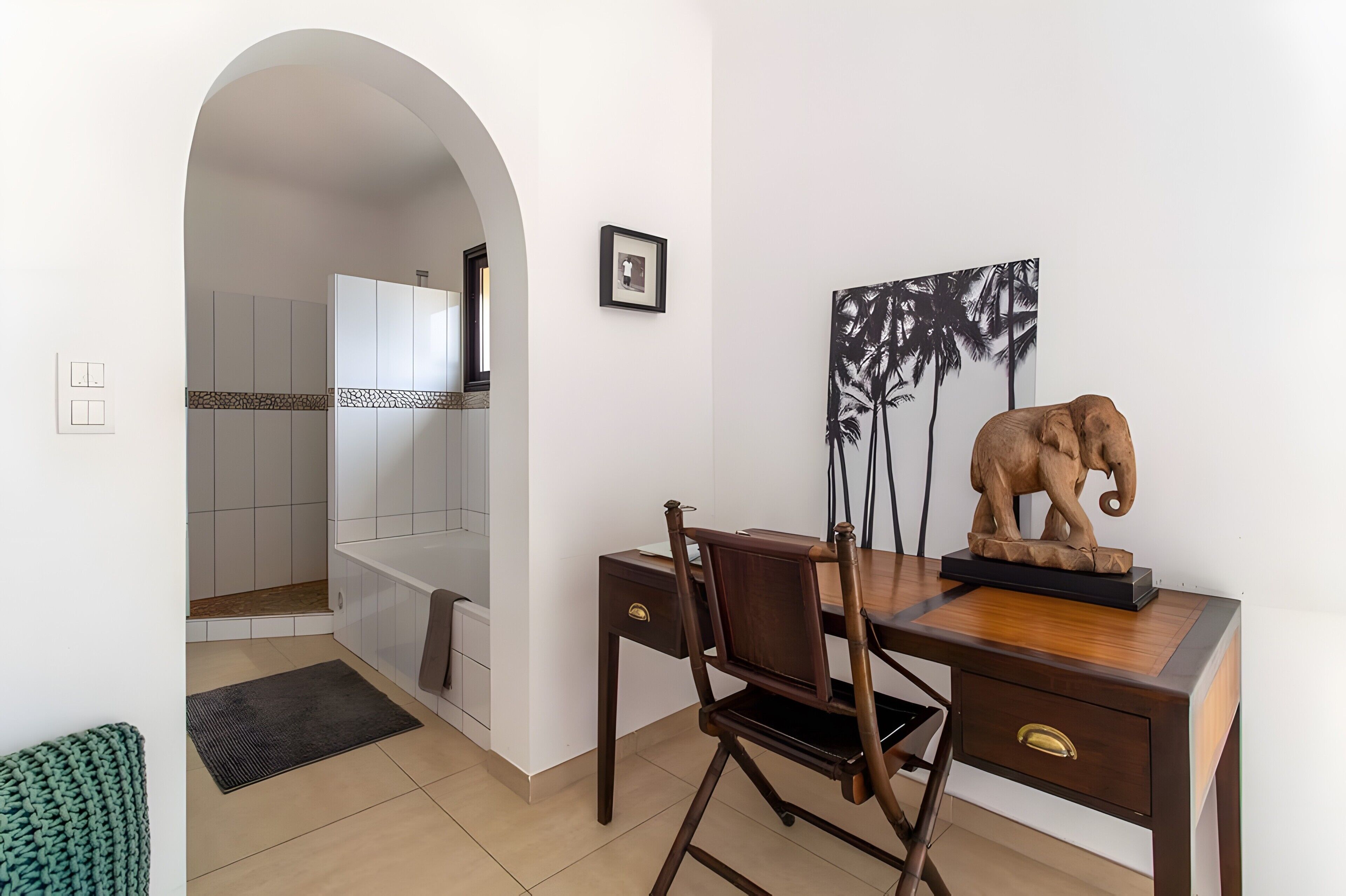 A room with a desk, chair, elephant sculpture, framed picture, and an archway leading to a shower area.