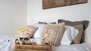 Un lit bien fait, agrémenté d’oreillers à motifs, un panier en osier contenant une tasse et un tableau accroché au mur.