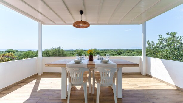 An outdoor dining area with a table set for two, white chairs, and a hanging light fixture. - Villa Francesco with Sea View (Orosei)