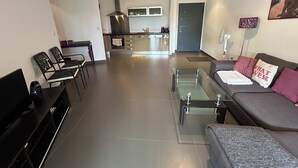 A modern living room with a sofa, a glass coffee table, and a TV. There is a kitchen area in the background with a stove and cabinets. - Between the upper town and the marina (Porto-Vecchio)