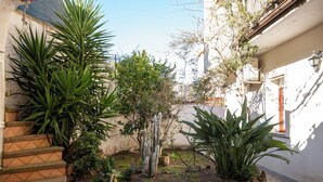 A courtyard with a variety of plants, a stone staircase, and a building with a white exterior and a red door. - House Gennauara (Lanusei)