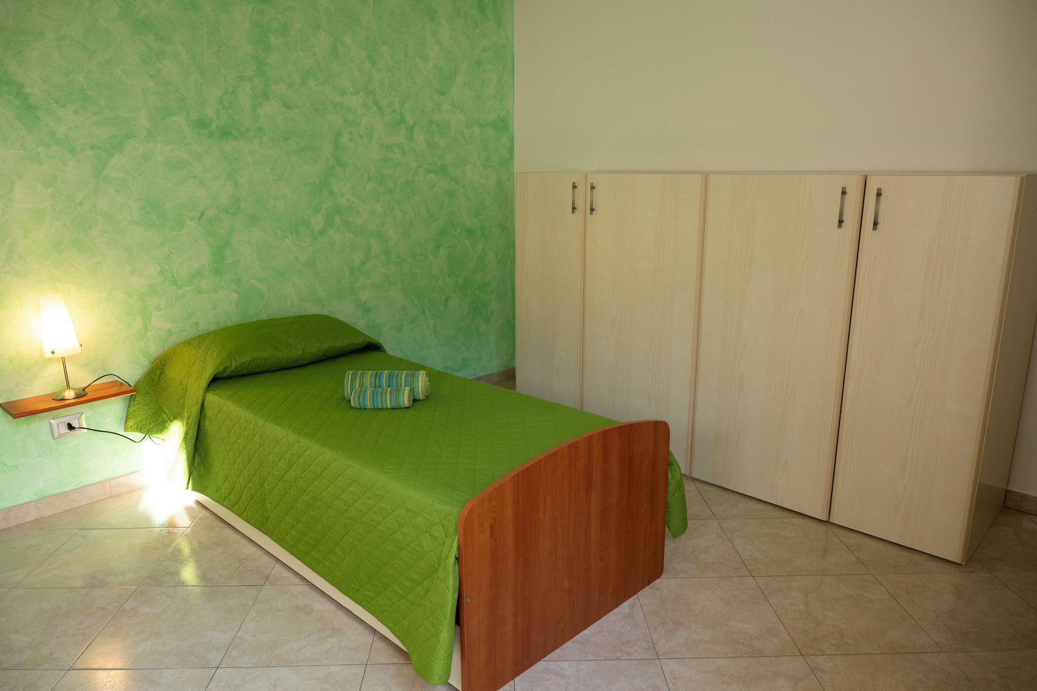 A bedroom with a green bed, a nightstand with a lamp, and a wardrobe.
