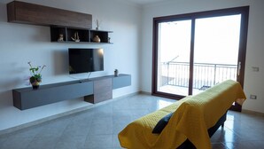 A modern living room with a flat-screen TV, a bench with a yellow cover, and a balcony with a railing. - House Gennauara (Lanusei)