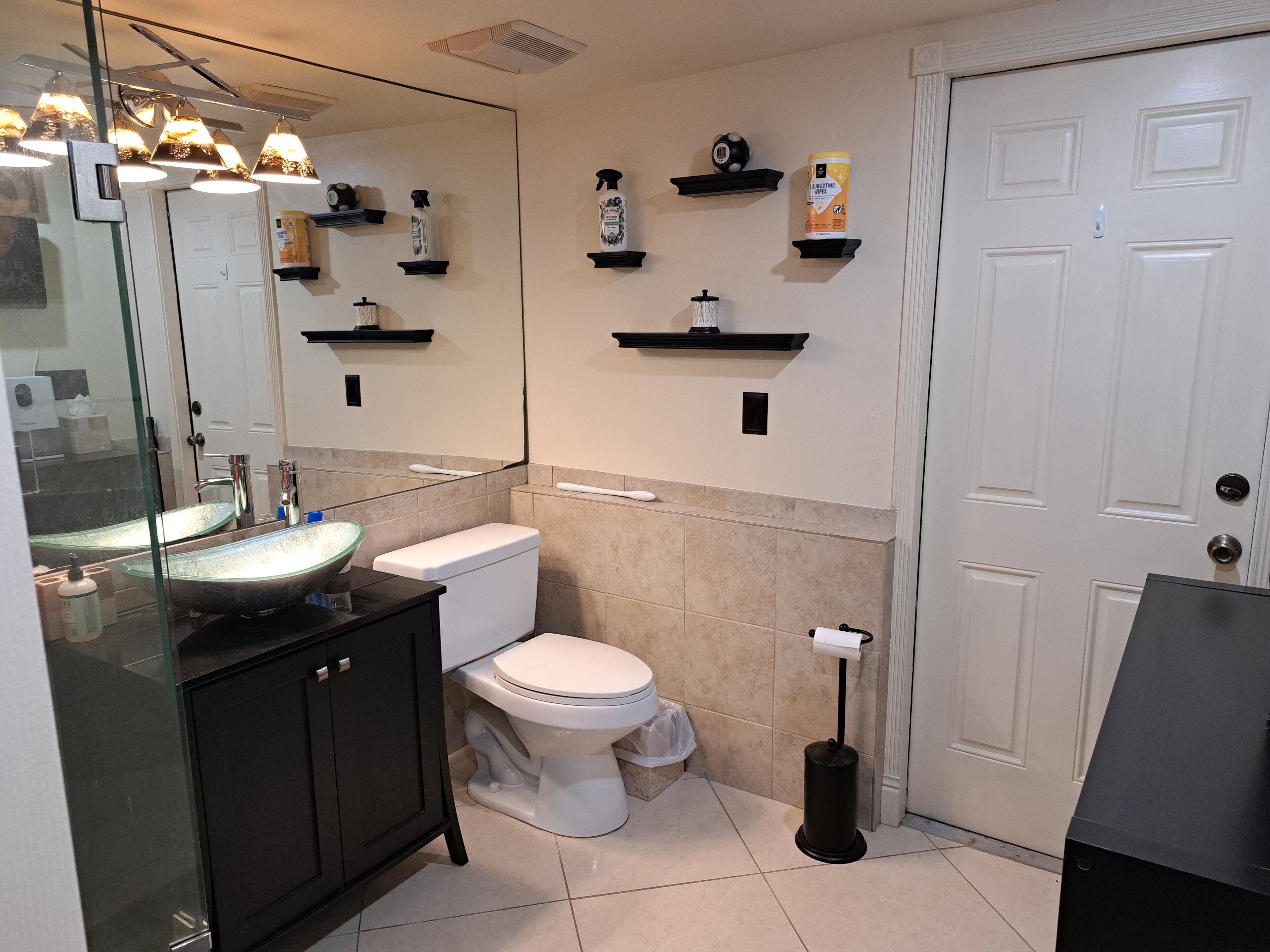A modern bathroom with a glass sink, white toilet, and beige tiles. There are shelves with toiletries and a mirror above the sink.