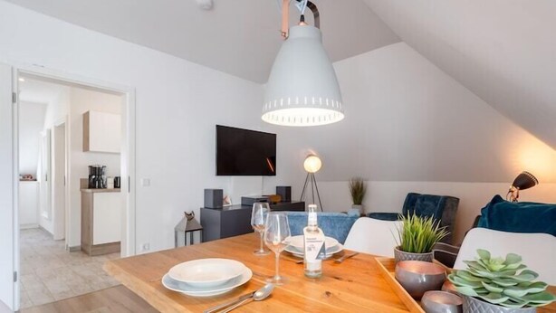 Un espace repas moderne avec une table en bois, des assiettes, des verres et une bouteille. Un luminaire est suspendu au plafond au-dessus de la table.