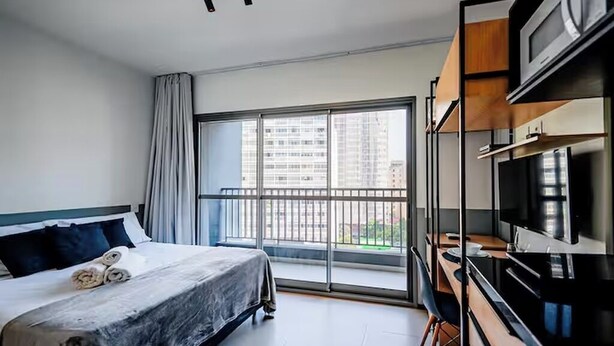 Een hotelkamer met een bed, een bureau, een televisie en een balkon met uitzicht op de stad.