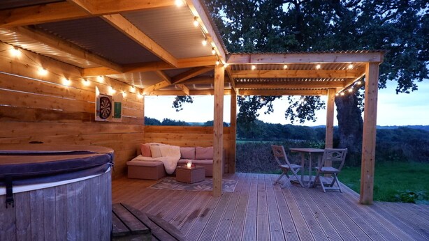 A wooden deck with a hot tub, seating area, and a pergola with string lights. - Shepherds Hideaway (Holsworthy)