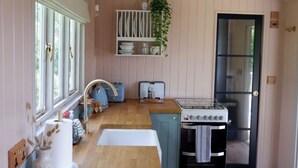 A kitchen with wooden countertops, a stove, and a refrigerator. There is a window with blinds, a plant hanging on the wall, and a shelf with dishes. - Shepherds Hideaway (Holsworthy)