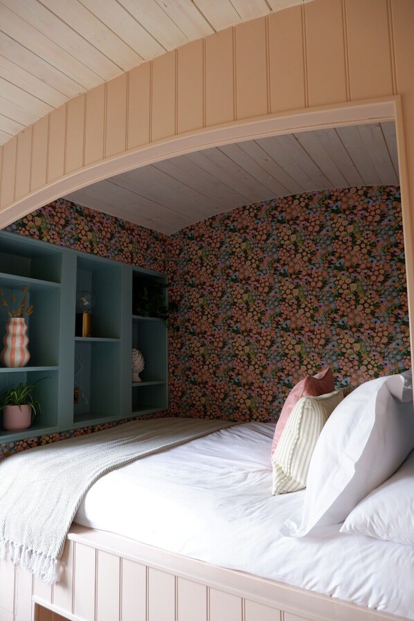 A bedroom with a bunk bed, floral wallpaper, a teal shelving unit, and a white bedspread. - Shepherds Hideaway (Holsworthy)