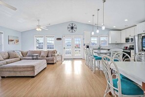 A spacious living room with a large sectional sofa, a dining area with white chairs, and a kitchen with white cabinets and stainless steel appliances. - Sea Why - Hot Tub - Ocean Views - Game Room (Surf City)