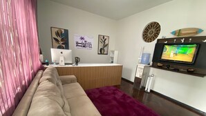 A living room with a beige sofa, a purple rug, a desk with a computer, and a TV mounted on the wall. - Açaizeiro Hostel (Belém)