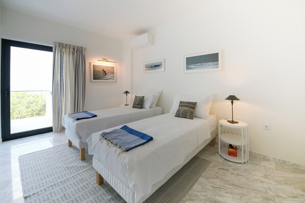 A bedroom with two beds, a window with curtains, a bedside table with a lamp, and a wall-mounted air conditioner.