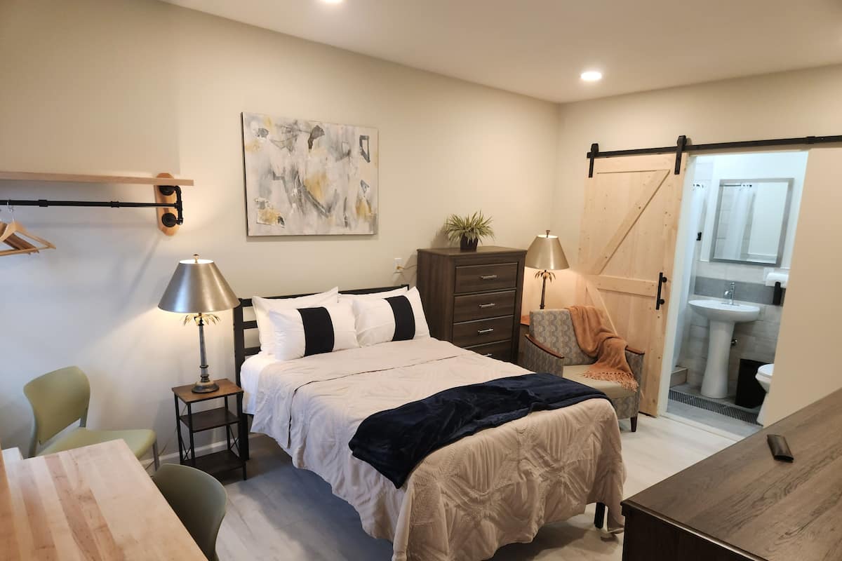 Une chambre moderne comprenant un lit, des tables de chevet, un bureau, une chaise, une armoire et une salle de bain visible à travers une porte ouverte.