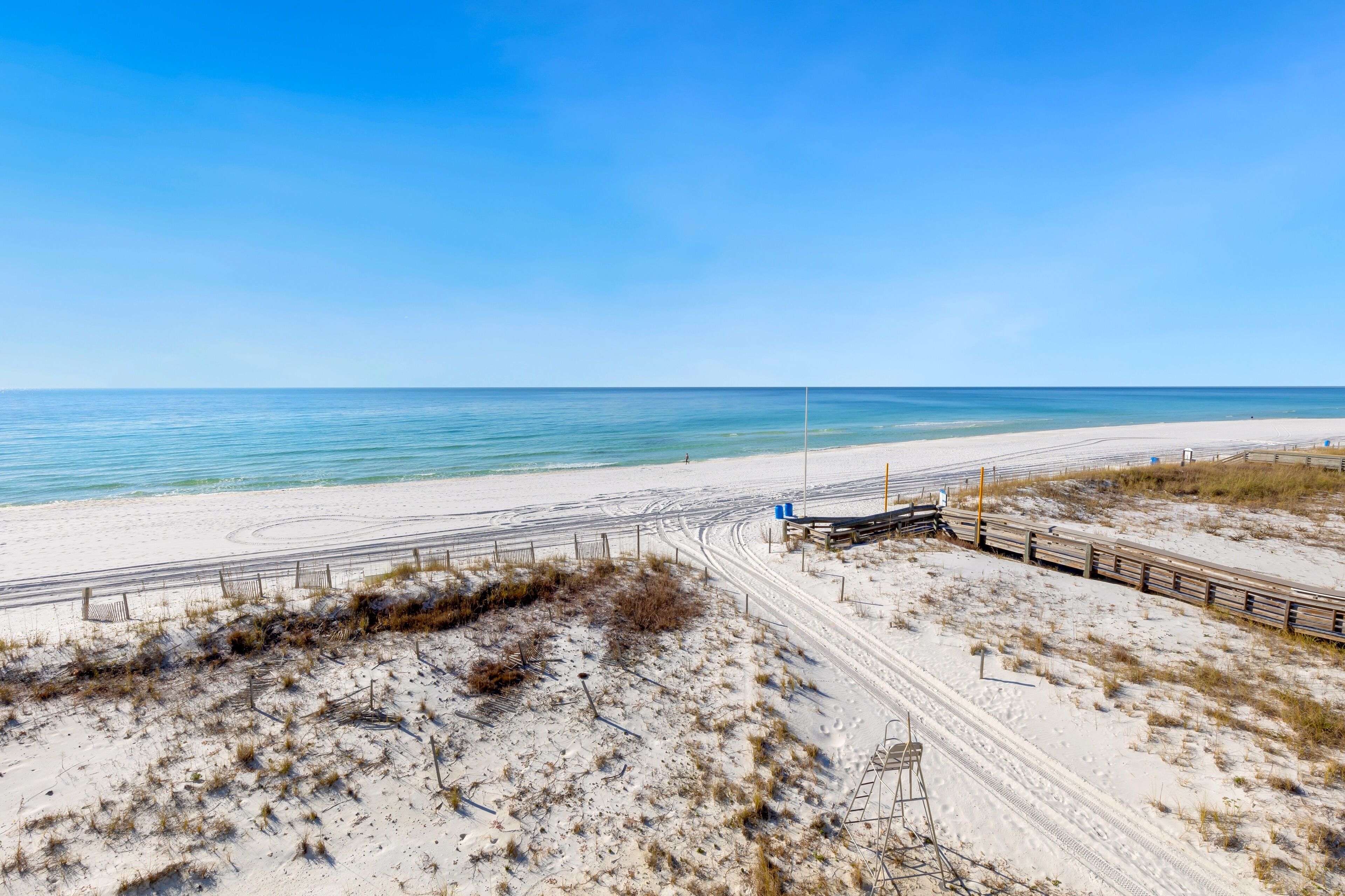 A beach with a sandy path leading to the water, a wooden fence, and a clear blue sky.