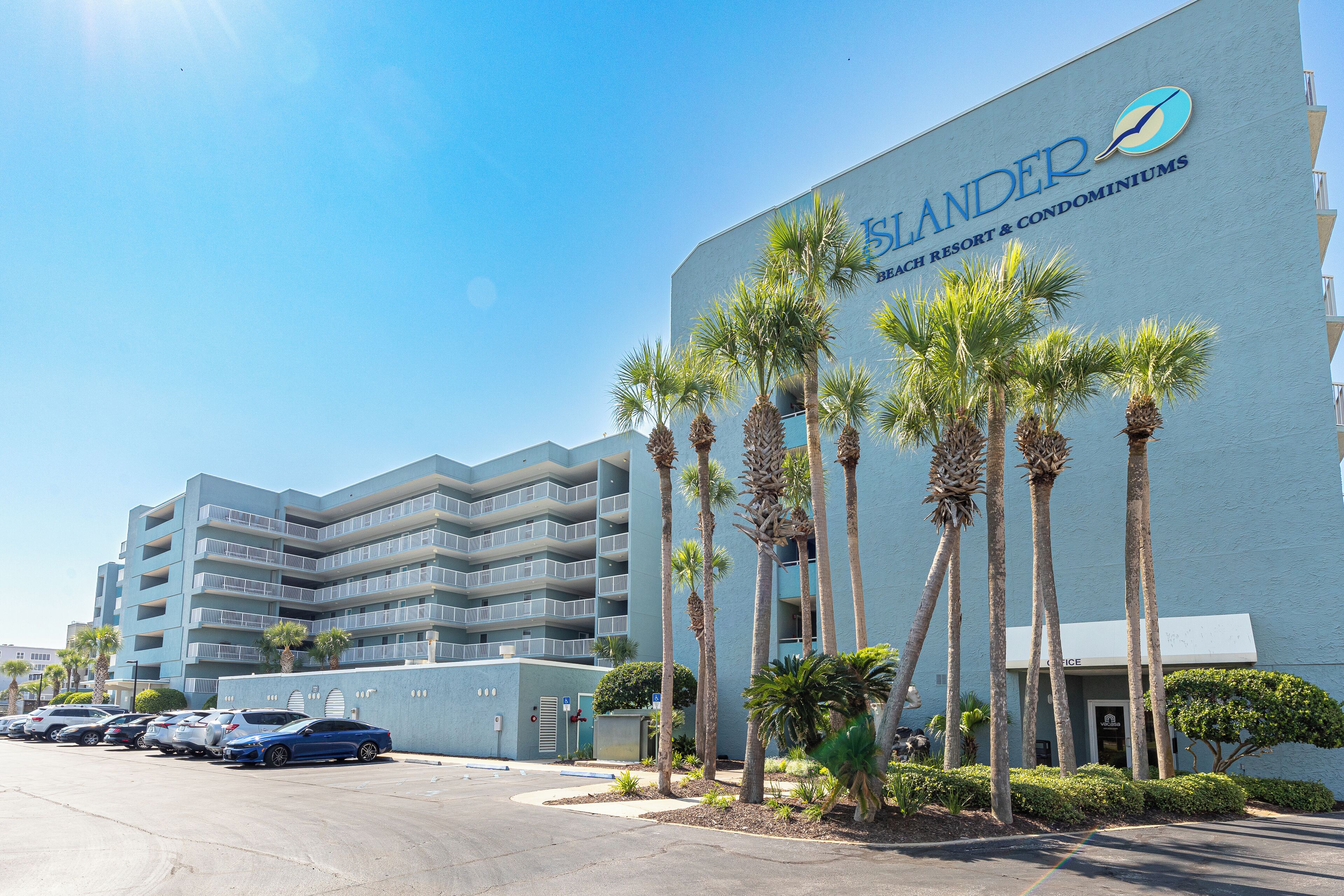 Ein Hotel direkt am Strand mit blauem Himmel, Palmen und einem Parkplatz.