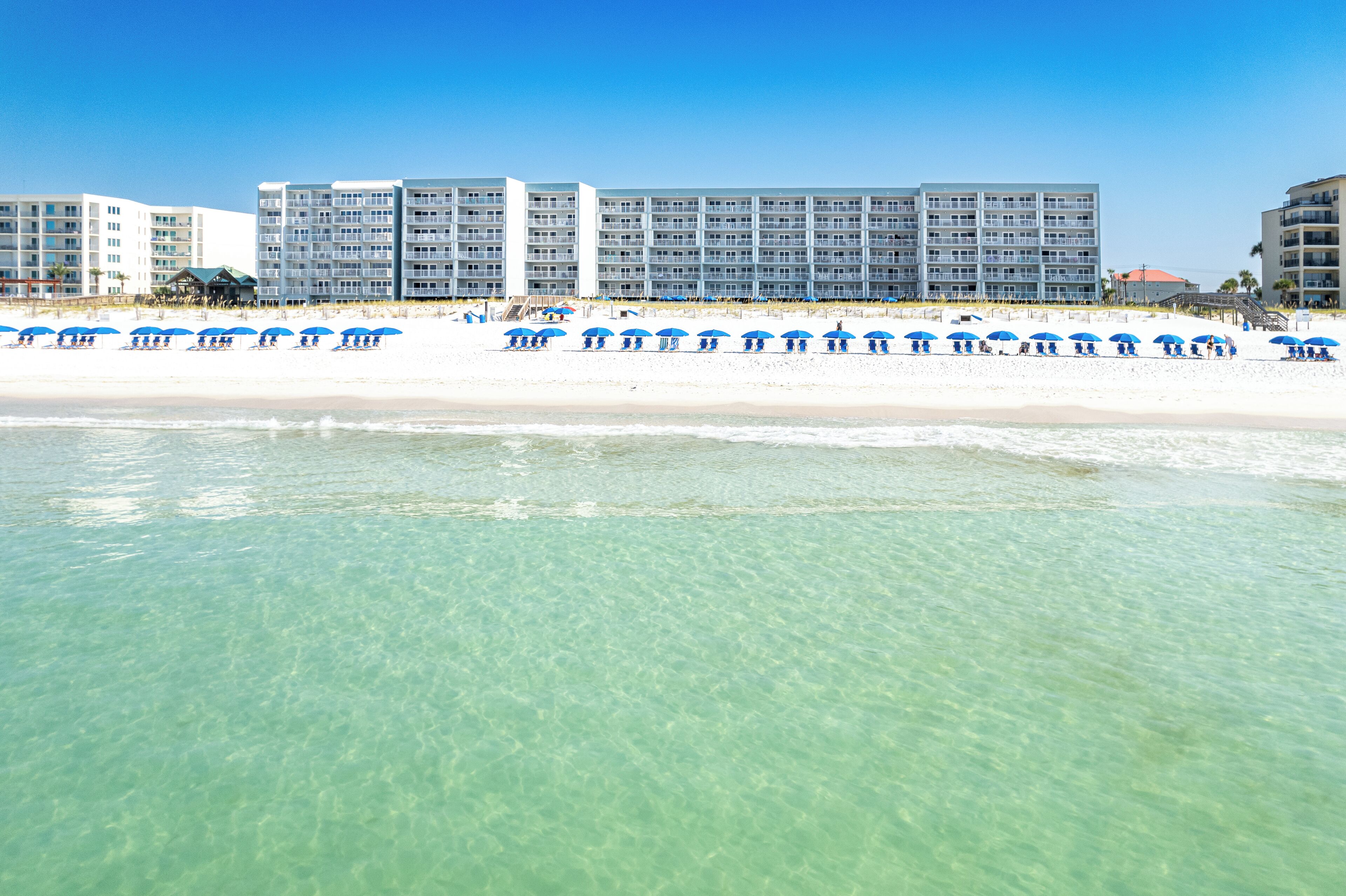 A beachfront hotel with multiple floors, a sandy beach, and blue umbrellas.