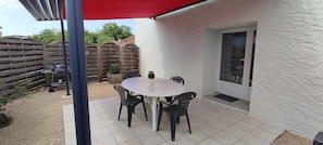 Une terrasse avec une table ronde et quatre chaises, un auvent rouge et un mur blanc à la texture particulière.