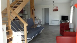 Un saló modern amb una escala de fusta, un sofà gris, un butaca vermella, una televisió i una zona de menjador al fons.