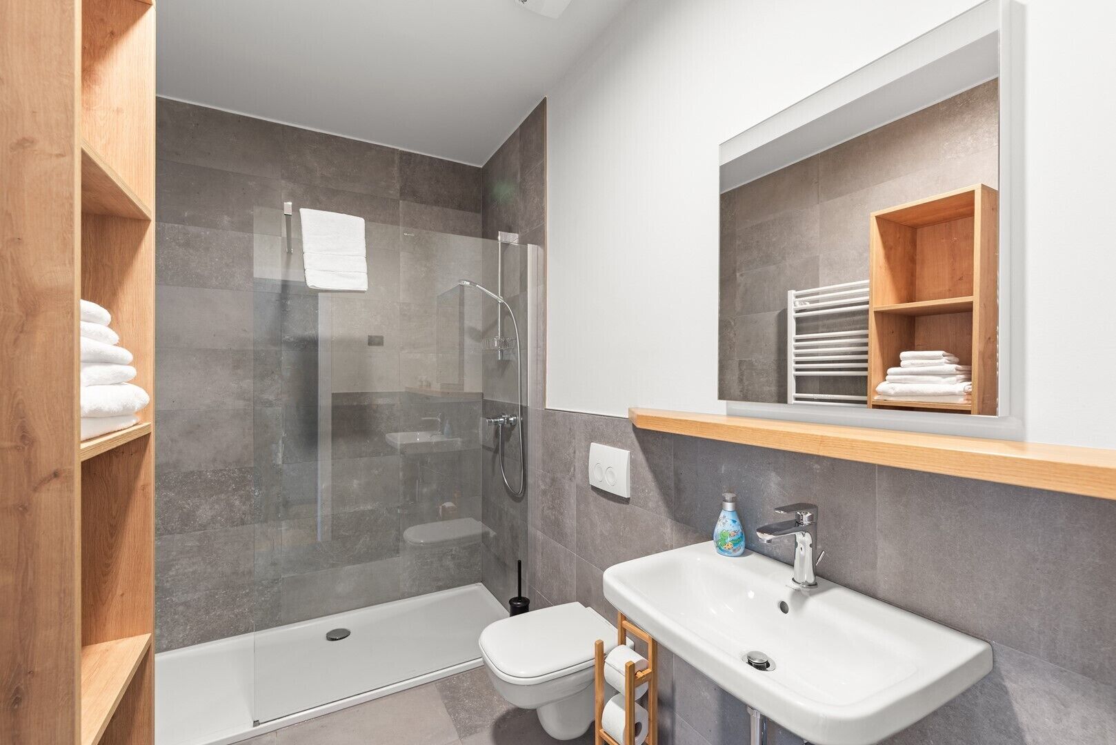 A modern bathroom with a glass-enclosed shower, a white sink, and a wooden shelf with towels.