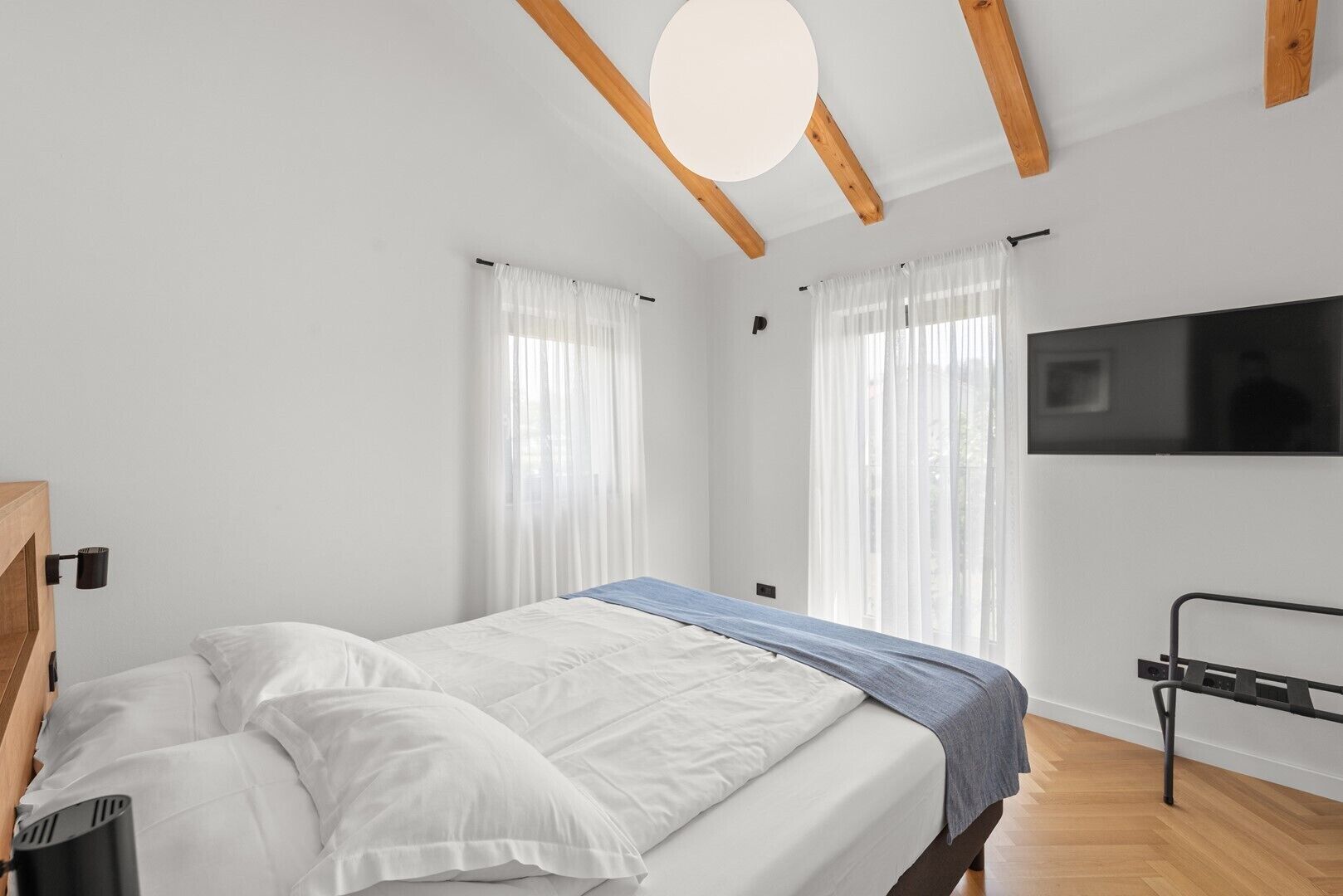 A modern bedroom with a bed, curtains, a TV, and wooden beams.